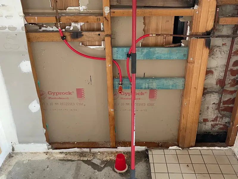 Opened wall showing new red water pipe and copper pipework above a tiled floor with a floor waste