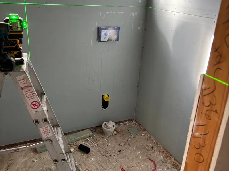 Bathroom walls lined with new wet-area board, a laser level on a stepladder and a toilet pipe outlet