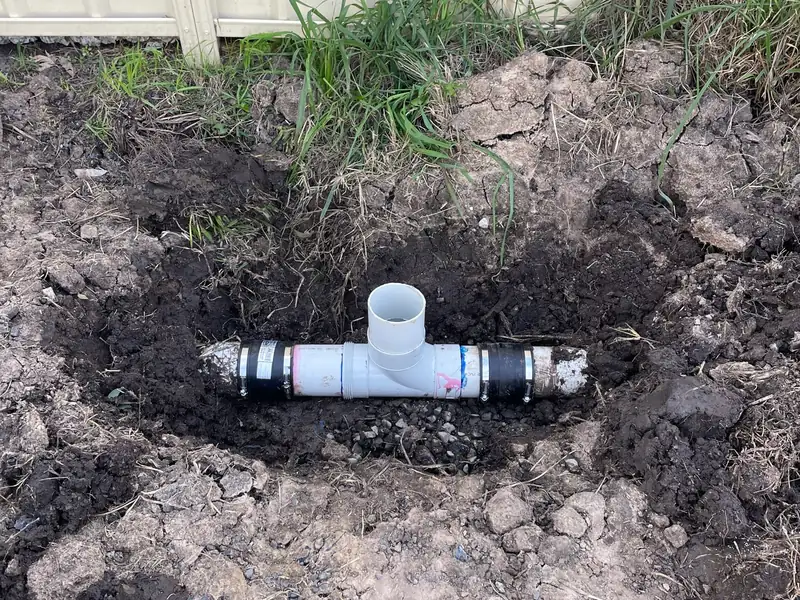 New PVC tee joined with rubber couplings in an excavated hole beside a steel fence
