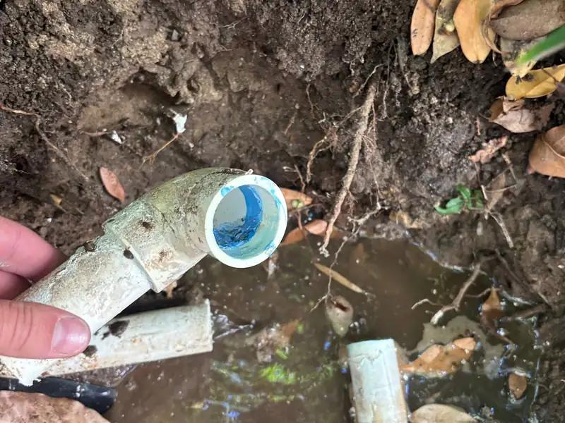 Broken PVC pipe elbow held up by hand over a muddy water-filled hole before repair