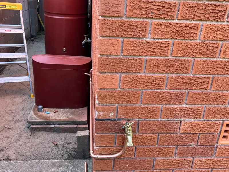 New copper pipe running down a brick wall to a brass garden tap beside two rainwater tanks