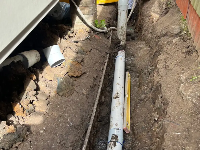 New PVC sewer line and riser bend in an open trench beside a house, spirit level alongside